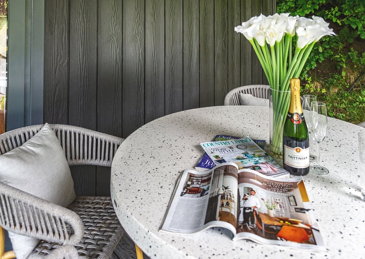 Exterior shot of a garden room with canopy close up shot of bistro table with bottle of champagne, glasses, open magazines and flowers in vase