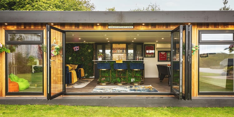Looking inside a luxury garden bar with bifold doors open so you can see bar seating and bright decor