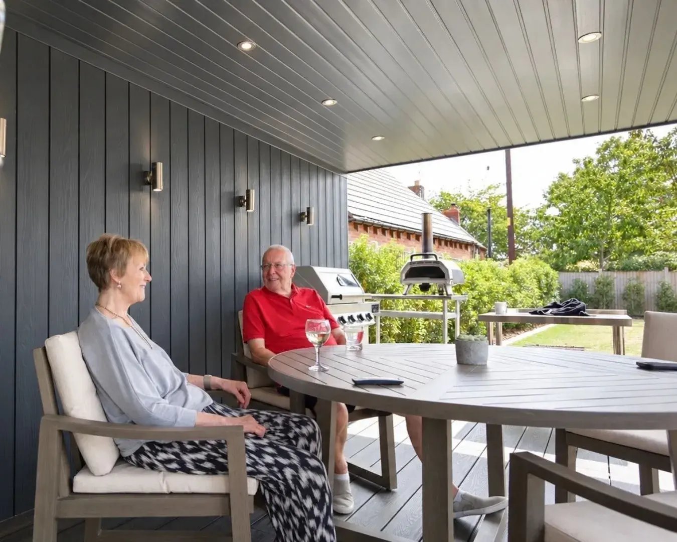 couple chatting at a dining table beneath their garden room’s covered veranda, with an outdoor kitchen.