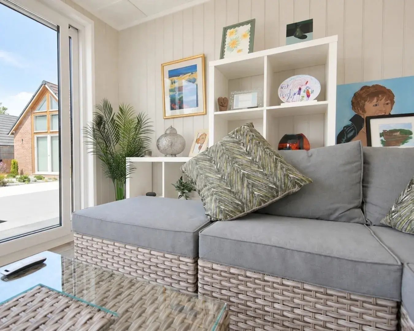 Garden sitting room interior with a grey rattan corner sofa, patterned cushions, artwork and glass doors overlooking the garden.