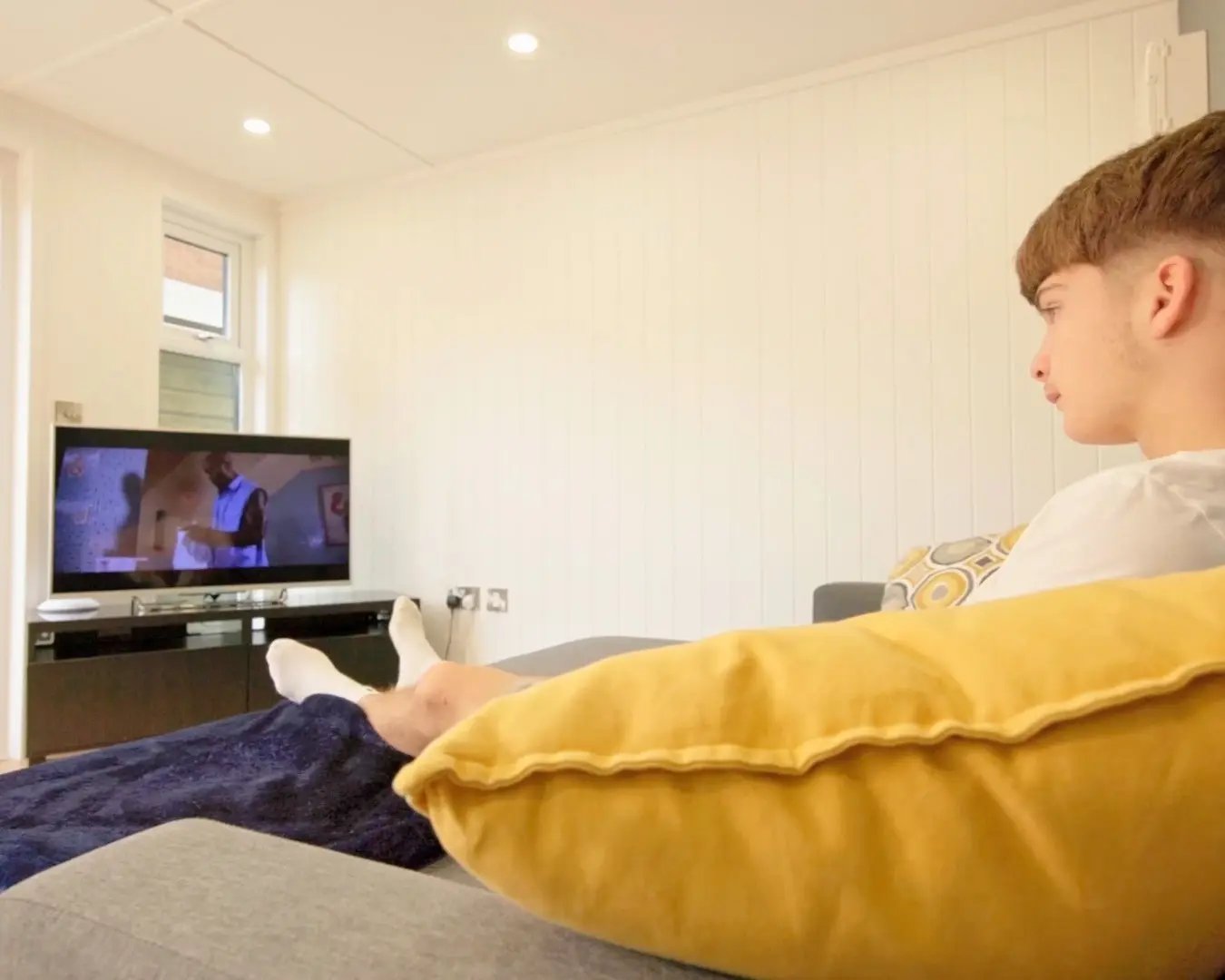 Teenager relaxing on a sofa and watching television inside a bright garden room den.