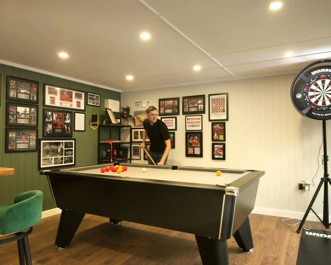 Man playing pool inside a modern garden room games room, with framed football memorabilia displayed on green and white panelled walls.