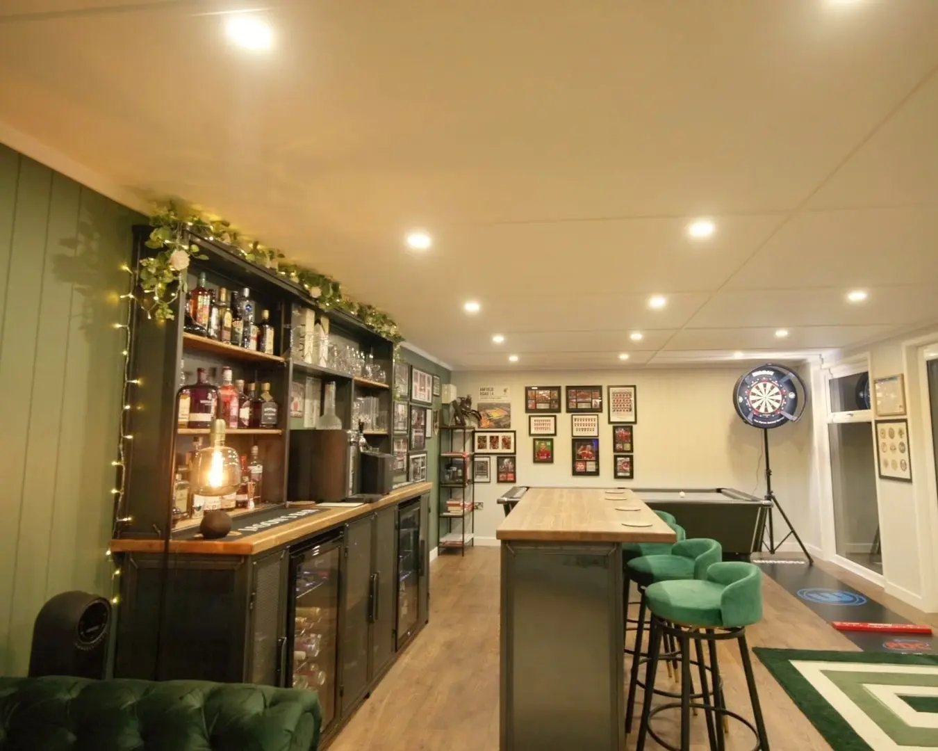 Spacious garden room interior with fitted bar, drinks shelving, pool table, dartboard, and wood-effect flooring, designed as a home entertainment space.