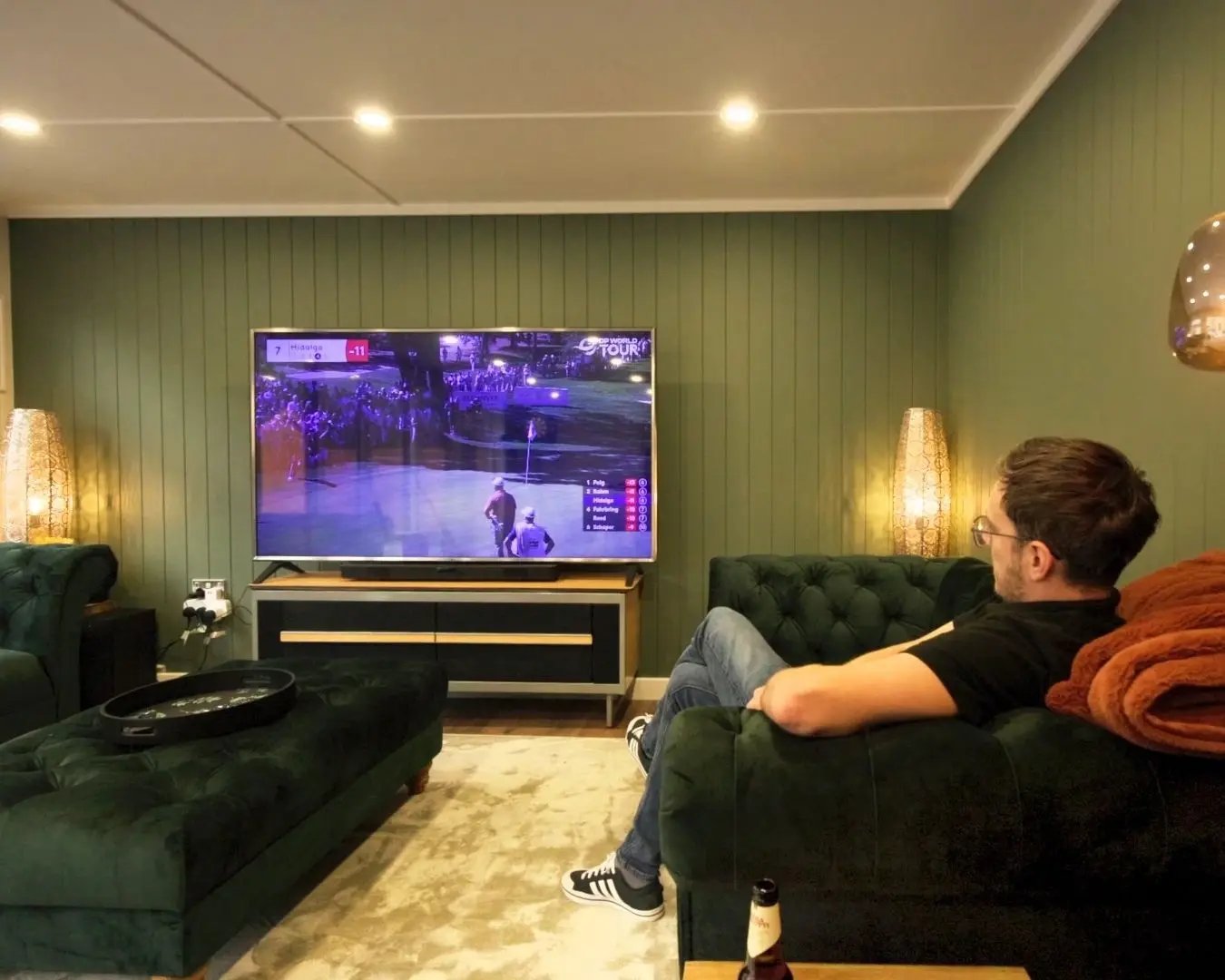 Man relaxing on a green velvet sofa inside a garden room, watching sport on a large television in a cosy lounge setting.