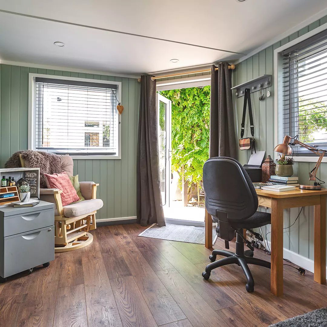 Internal shot of Cabin Master Small Garden Office Pod with desk, lamp, book and magazines in foreground and furry brown armchair and carpet in background