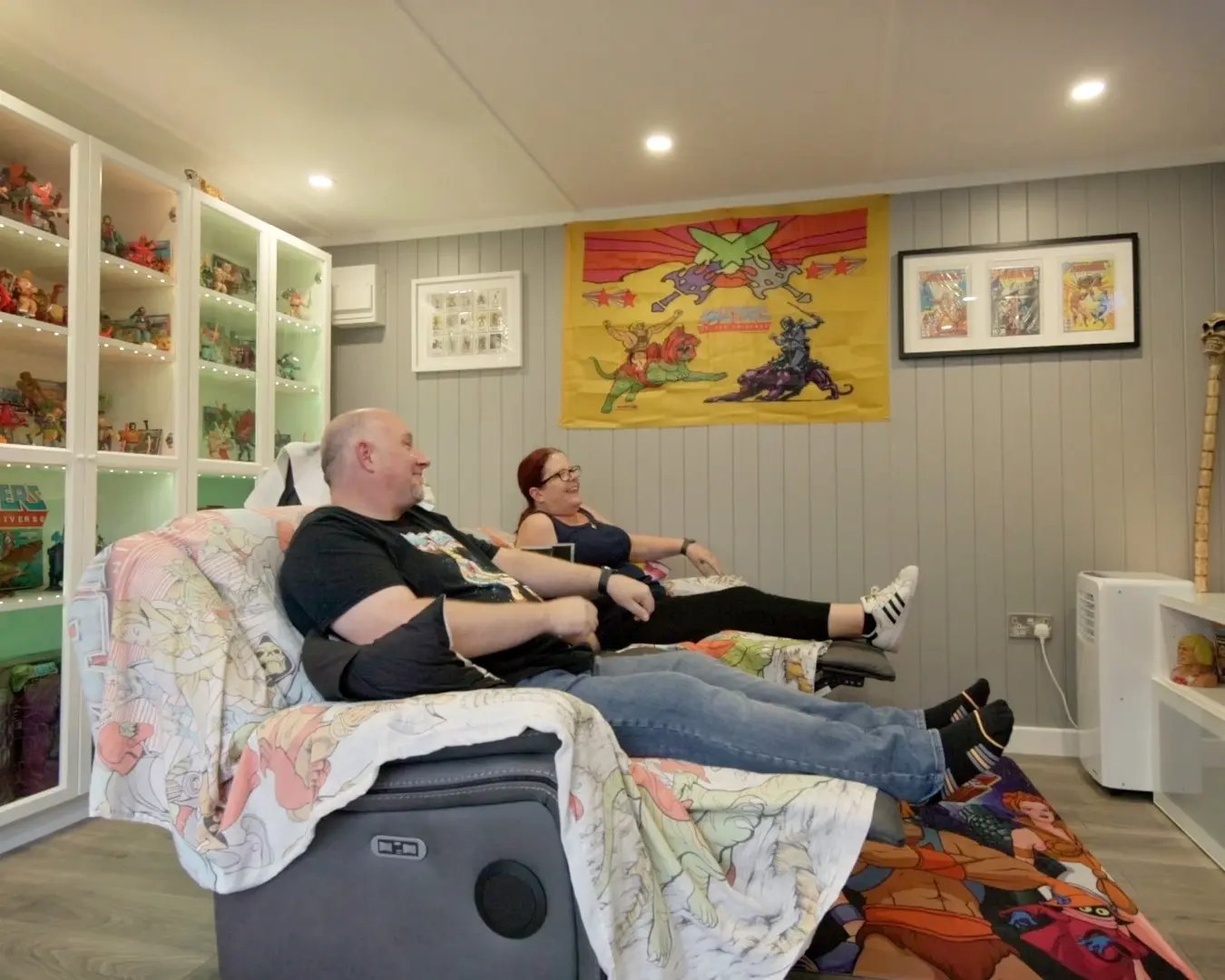 Couple in garden room watching Tv together sitting on sofa surrounded by he man collection