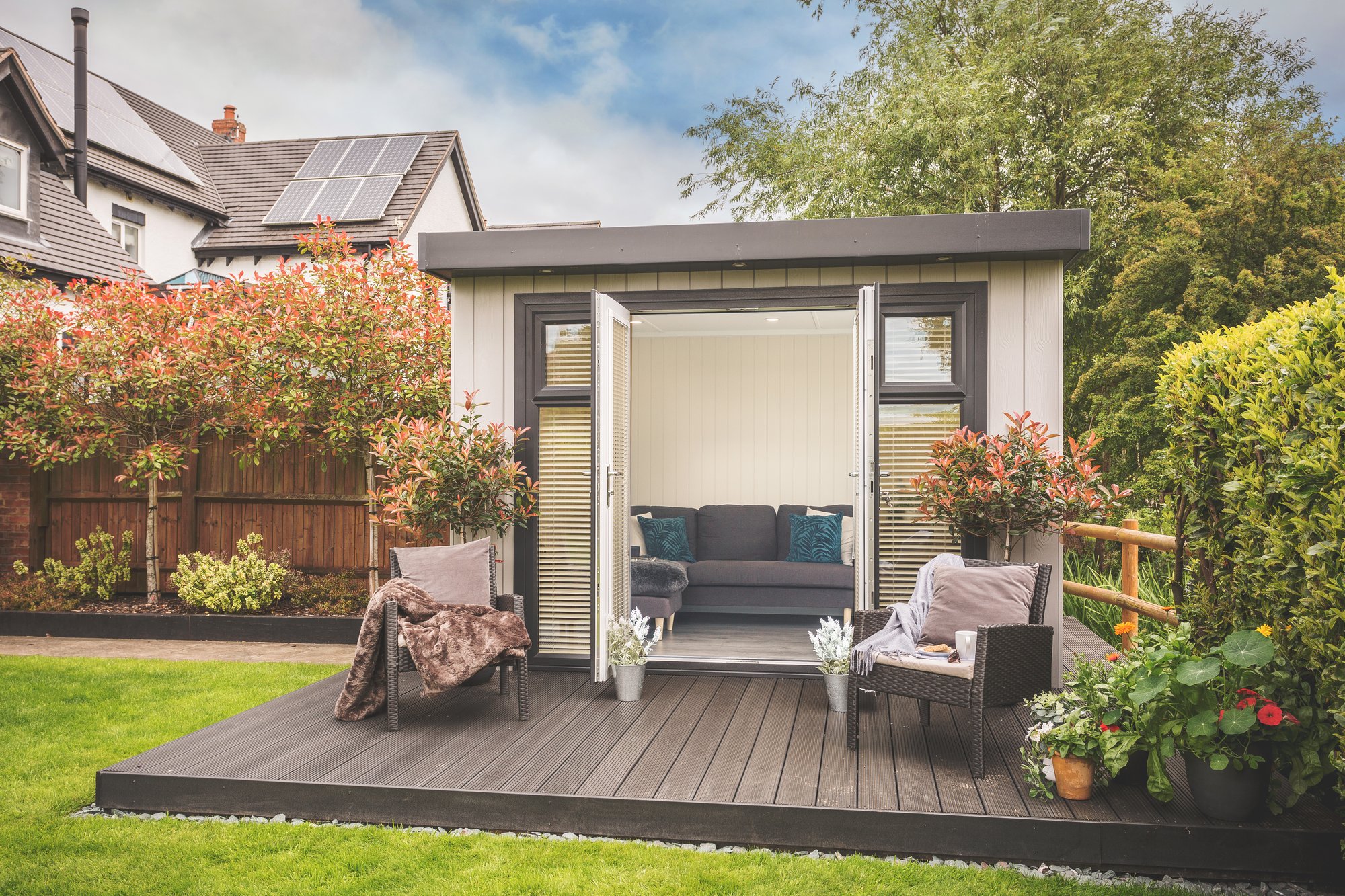 Small garden summerhouse with large timber decking in front and 2 rattan armchairs with throws on top of decking.