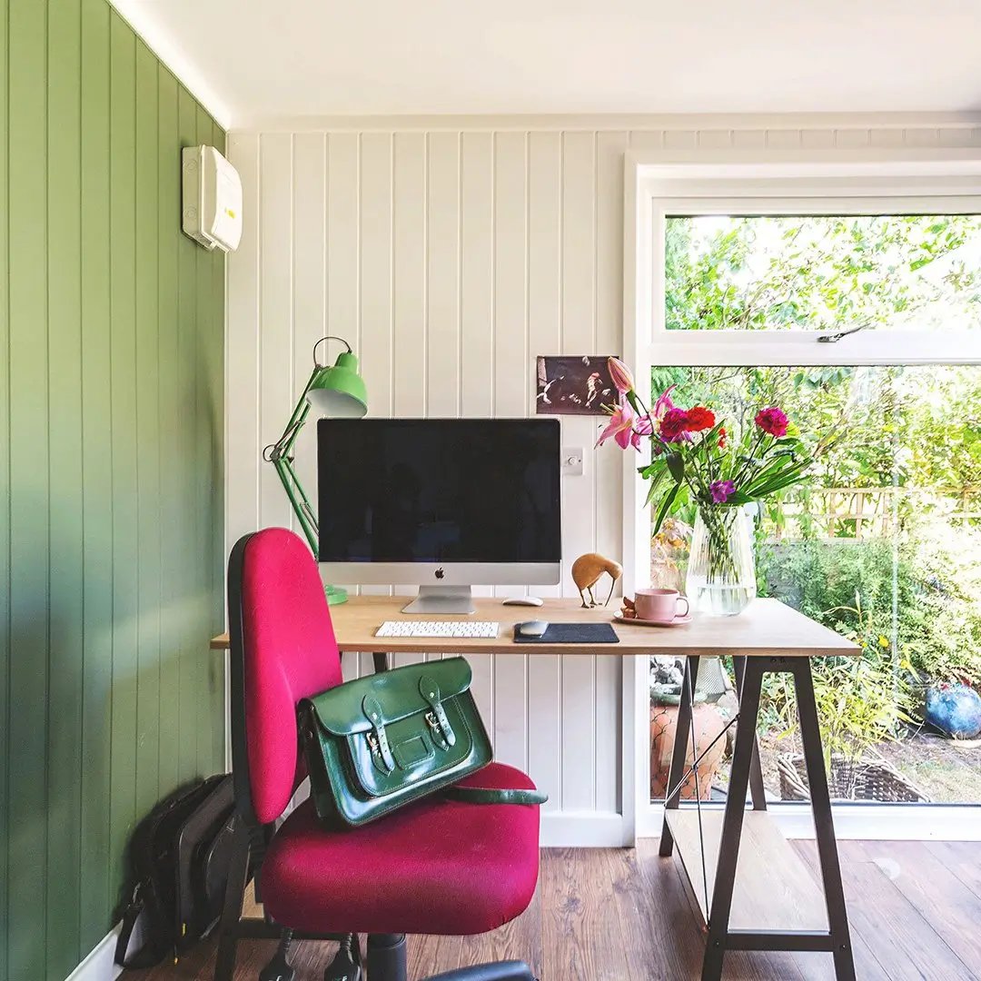 small garden office with pink desk chair and mac computer with patent leather green satchel and vase of flowers on top of it 