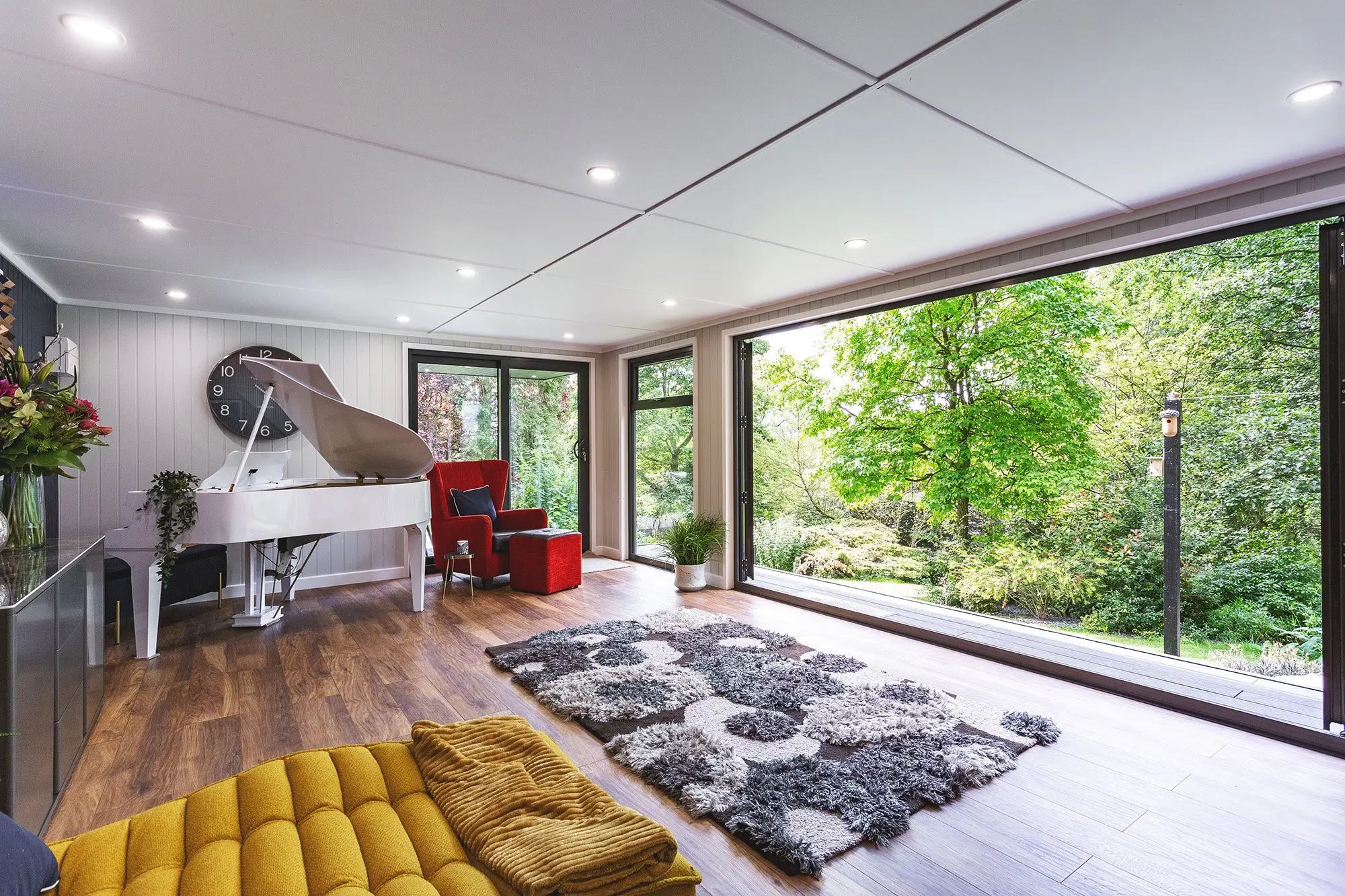 Large marley veranda garden room with large open bifold doors onto grassy lawn area with monochrome rug and large white piano in corner of the room