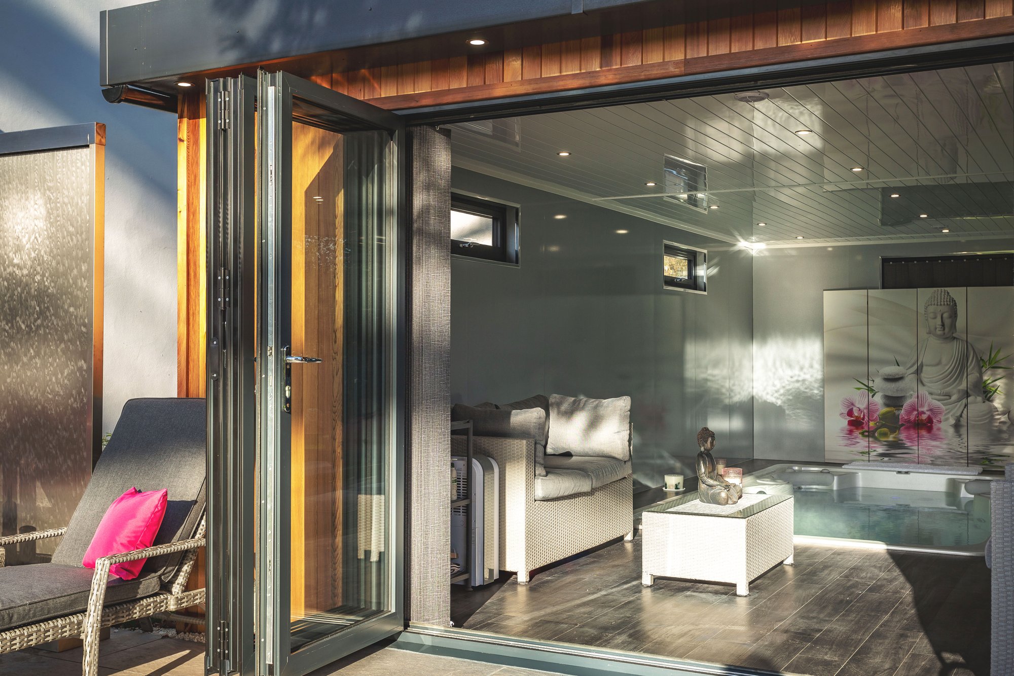 Large swim spa garden building with open bifold doors and white rattan sofa and table with a buddha statue on top of it