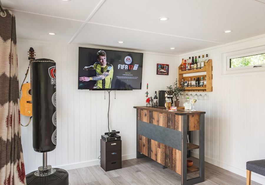 Bespoke garden games room featuring a rustic wooden bar, wall-mounted TV, freestanding punch bag and white panelled walls.