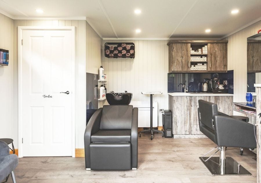 Bespoke garden hair salon featuring a black hair-washing chair, styling station, storage cupboards and a separate toilet.