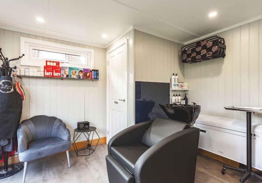 Modern garden hair salon with a reclining wash chair, black shampoo basin, product shelves and a comfortable waiting area.