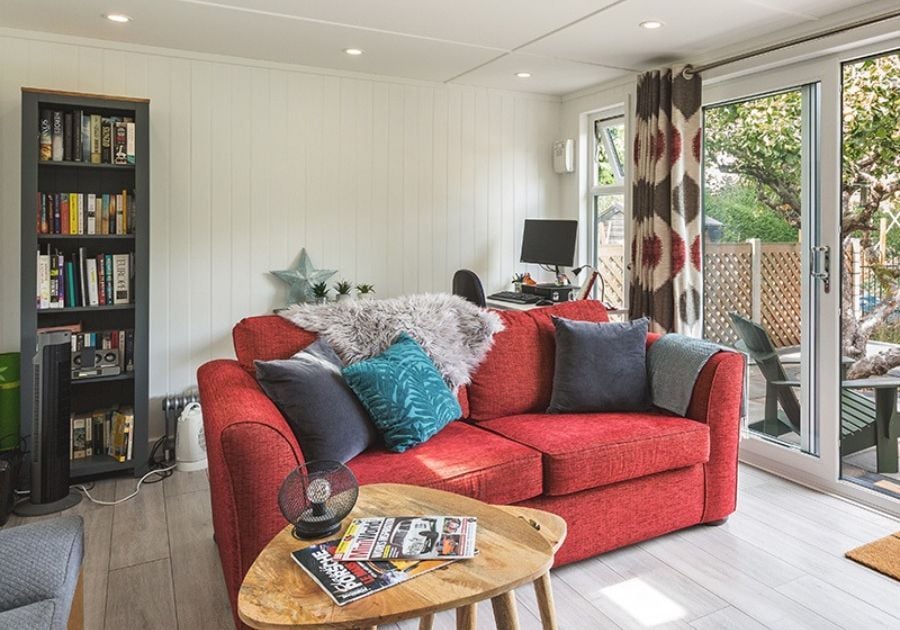 Cosy garden room lounge featuring a red sofa, wooden coffee table, bookcase and computer desk with glazed doors overlooking the garden.