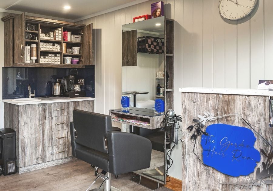 Garden hair salon featuring a professional styling chair, large mirror, hairdressing equipment, storage cupboards and reception counter.
