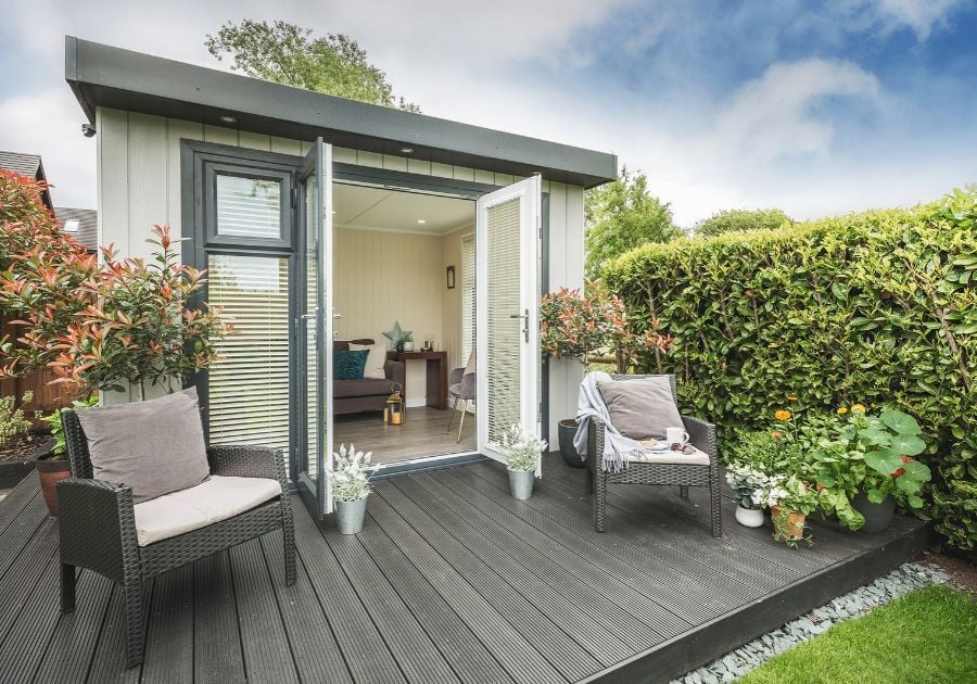 Modern grey-clad garden room with glazed double doors, outdoor decking, wicker seating and potted plants surrounded by greenery.