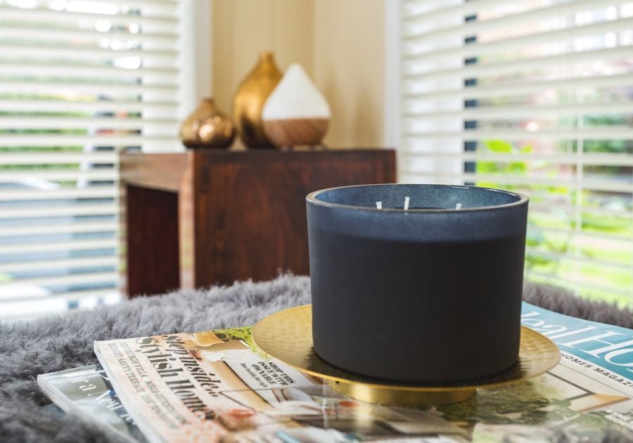 Decorative candle displayed on a glass table with magazines and a soft grey throw inside a cosy garden room with white Venetian blinds.