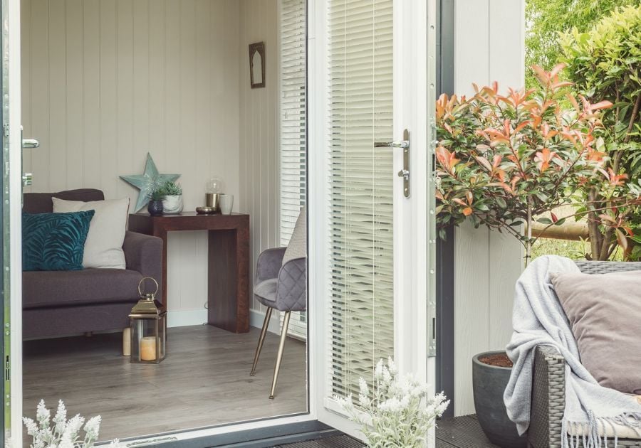 Cosy garden room lounge featuring a grey sofa, teal cushions, decorative furnishings and glazed double doors opening onto the garden.