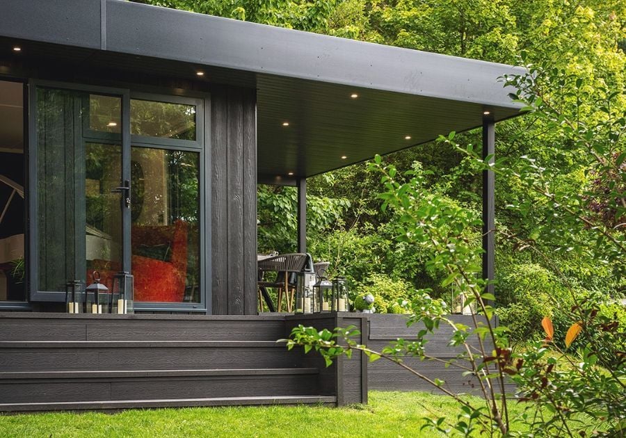 Contemporary garden room with black composite timber cladding, a covered outdoor seating area, decking and large windows surrounded by greenery.