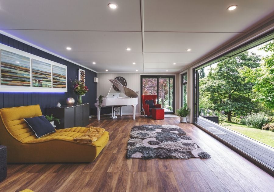 Bespoke garden music room featuring a white grand piano, mustard yellow chaise longue, modern furnishings and floor-to-ceiling glazing overlooking the garden.