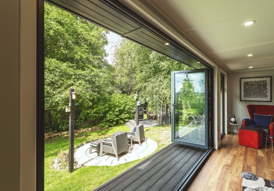 Modern garden room with large open glass doors leading onto grey decking and a landscaped garden with an outdoor seating area.
