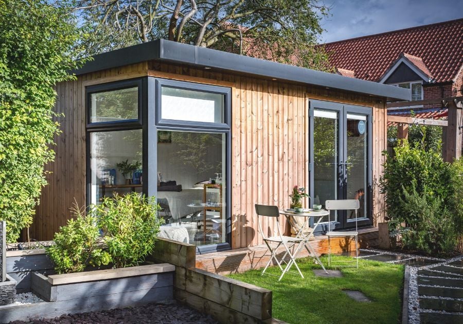 Bespoke redwood -clad garden beauty salon with anthracite grey double doors, large corner windows and a small outdoor seating area.