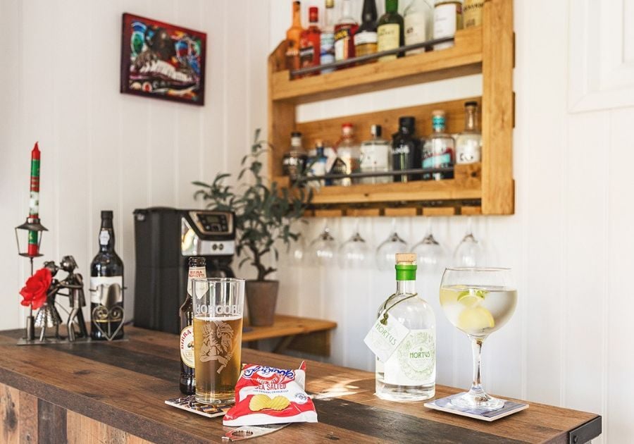 Rustic wooden garden bar with wall-mounted drinks shelves, hanging glasses and a selection of drinks inside a bespoke garden room.