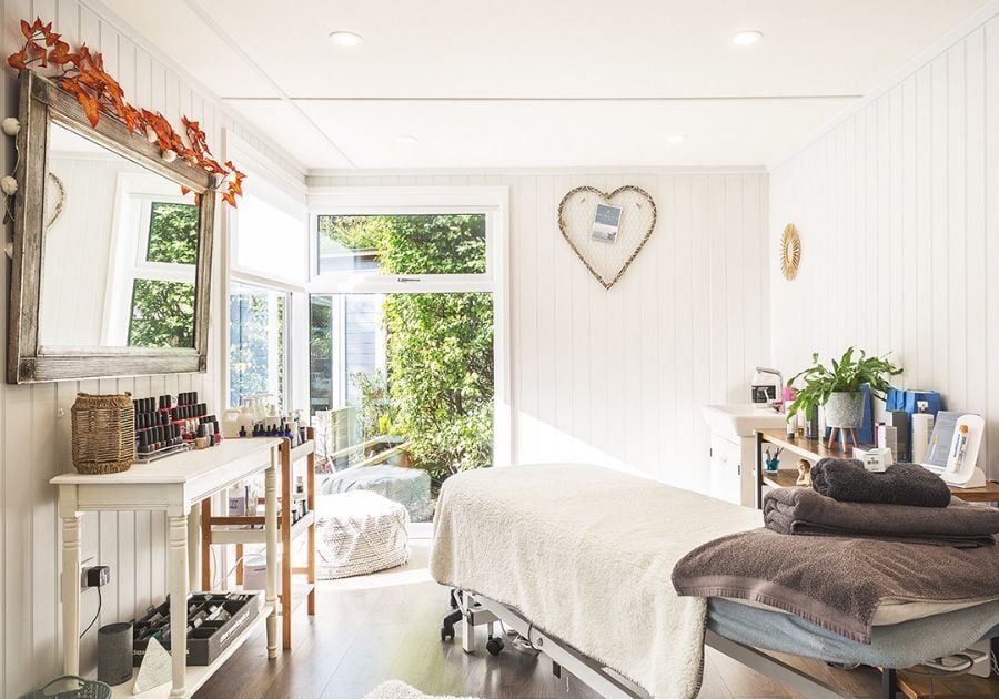 Bright garden beauty salon featuring a treatment bed, nail polish display, rustic wall mirror and large windows overlooking the garden.