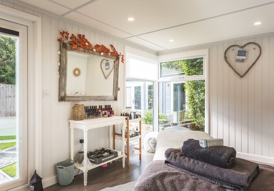 Modern garden beauty salon featuring a massage treatment bed, beauty products, white panelled walls and large windows providing natural light.
