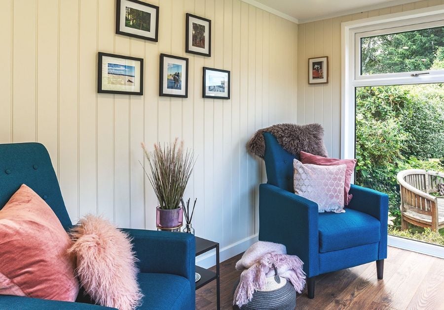 Cosy garden room seating area featuring two blue armchairs, pink cushions, framed wall art and large windows overlooking the garden.