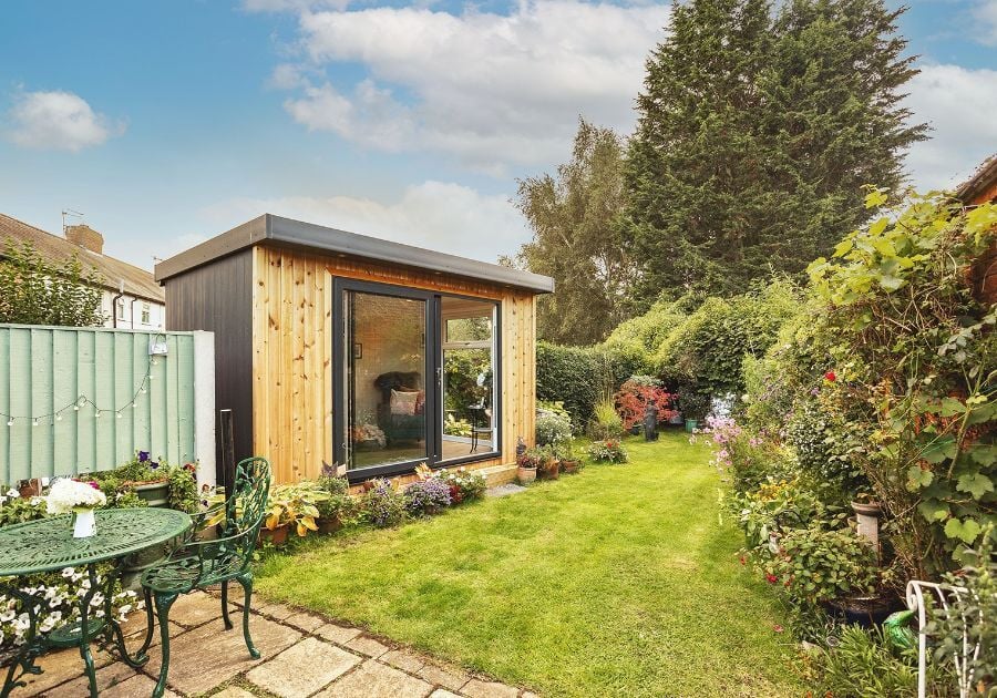 Modern timber-clad garden room with large sliding glass doors, positioned at the end of a landscaped garden with colourful flower beds and patio seating.