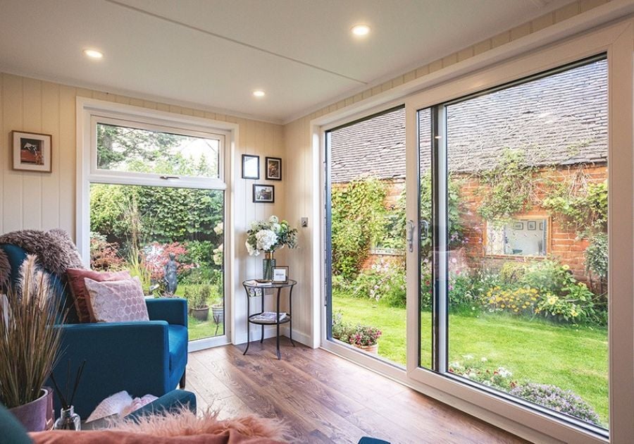 Bright garden room lounge featuring blue armchairs, pink cushions, wooden flooring and large sliding glass doors overlooking a colourful garden.