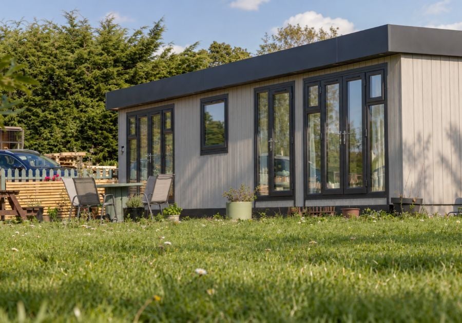 Contemporary garden annexe with grey cladding, anthracite grey glazed double doors and a flat roof, surrounded by a lawn and outdoor seating.
