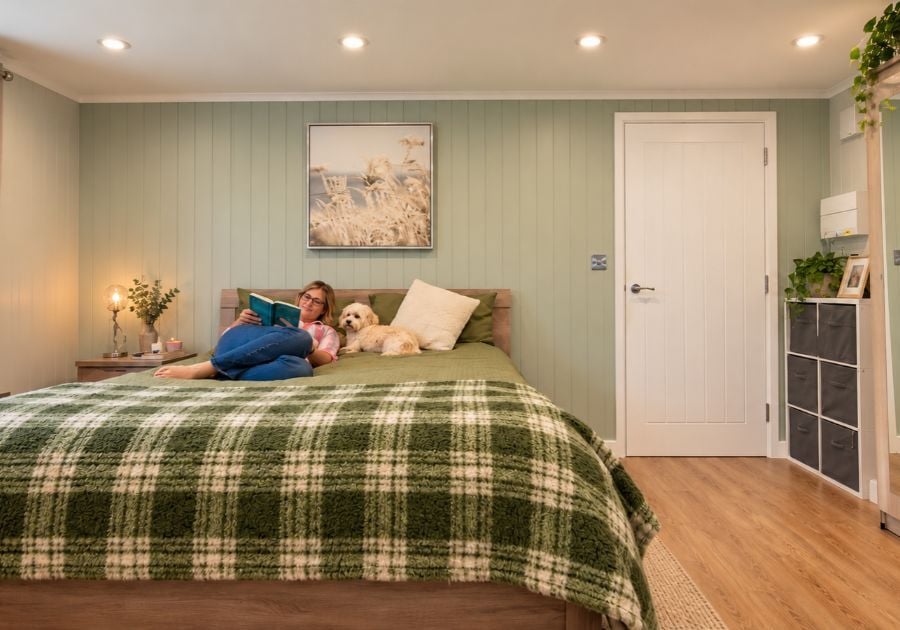 Cosy garden annexe bedroom featuring a double bed with green bedding, sage green panelled walls and a woman relaxing with her dog.