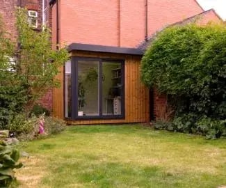 A small modern garden office with wooden cladding and large glass sliding doors, positioned in the corner of a neatly maintained garden beside a red-brick house. The office interior shows shelving and a desk, surrounded by green bushes and a lawn
