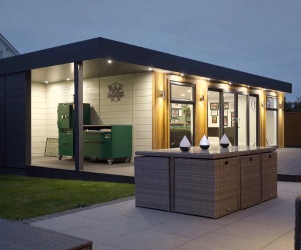 Modern garden room with veranda and outdoor kitchen area, featuring wood cladding, large sliding glass doors, and patio seating in a landscaped garden at dusk.
