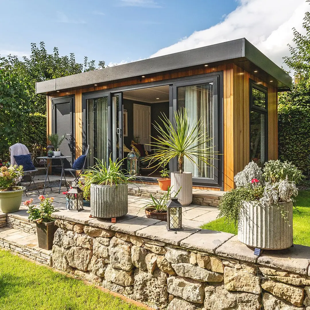 Small Garden Room Summerhouse in patio area with potted plants and large open sliding glass doors with garden furniture in front of it 