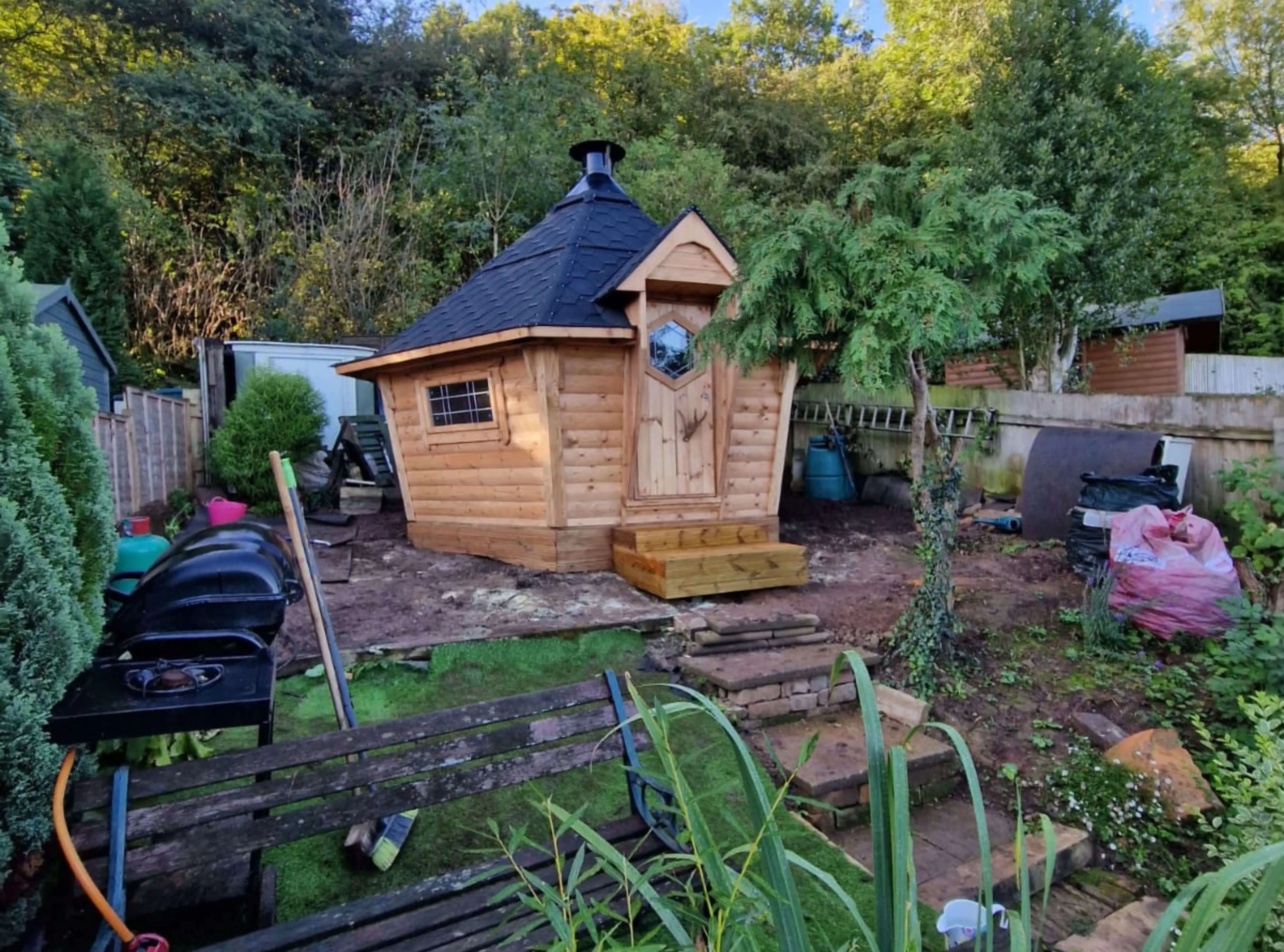 BBQ hut with black roof in garden with green trees and concrete steps and mud and plants and outdoor BBQ and hose pipe 