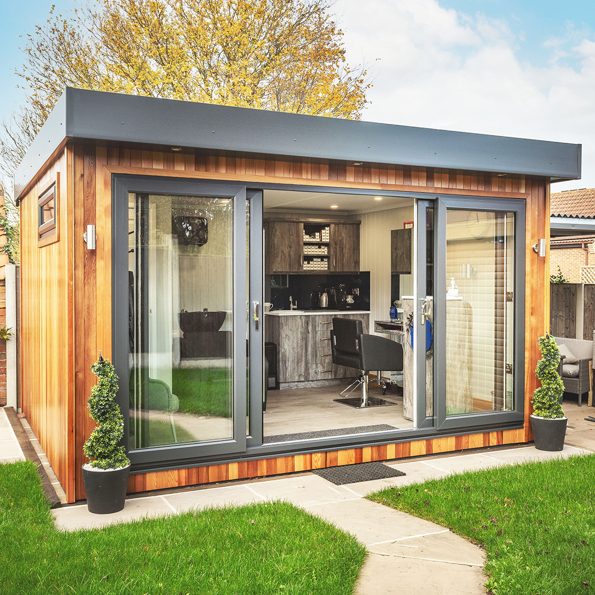 Cabin Master Hairdresser Studio in garden with patio pathway and grassy area and potted plants and sliding doors with internal view of room with black leather chair  