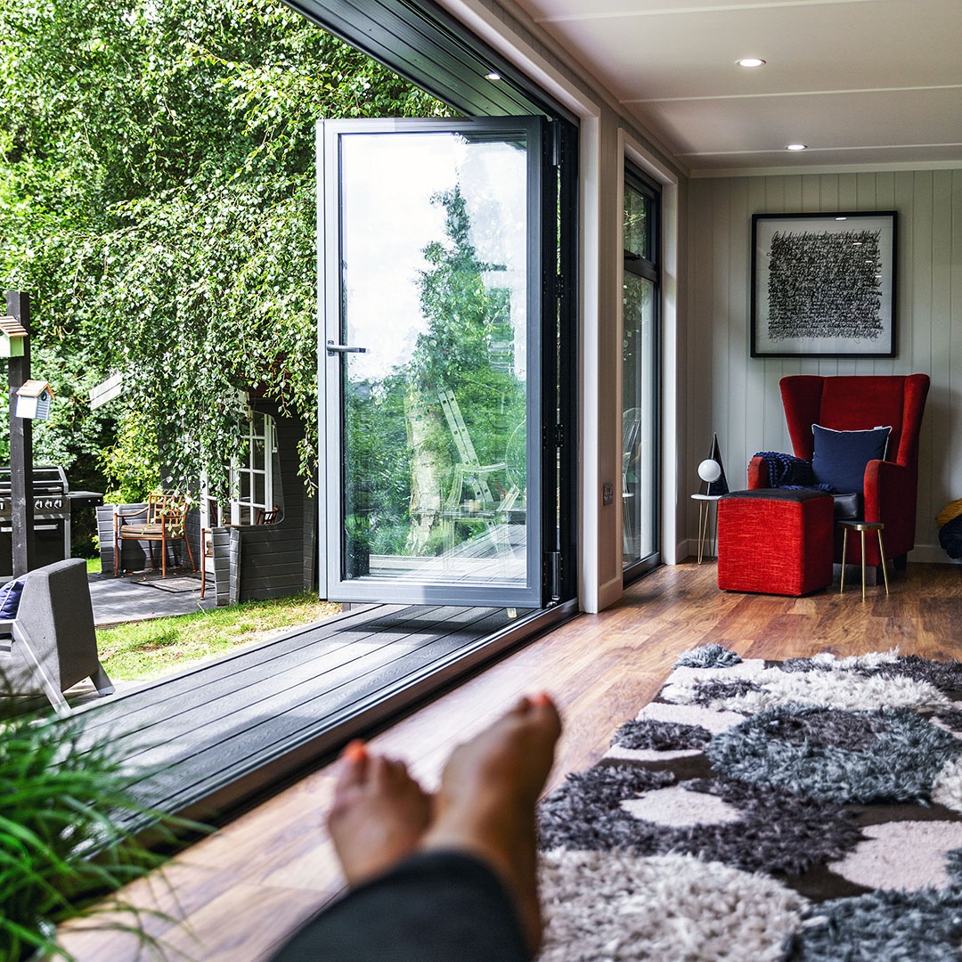 Internal view of cabin master garden room with composite decking and bi-fold doors and view of woman's feet in foreground with large rug and red velvet armchair