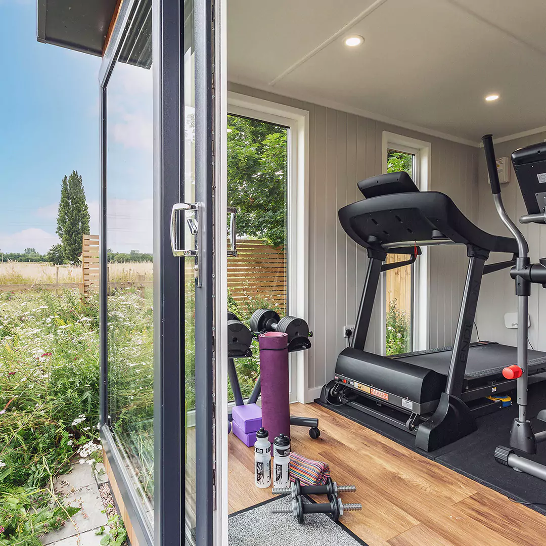 Cabin Master Garden Gym Building with sliding doors and view of running machine, dumbbells, mat and body roller with garden grass and flowers in background
