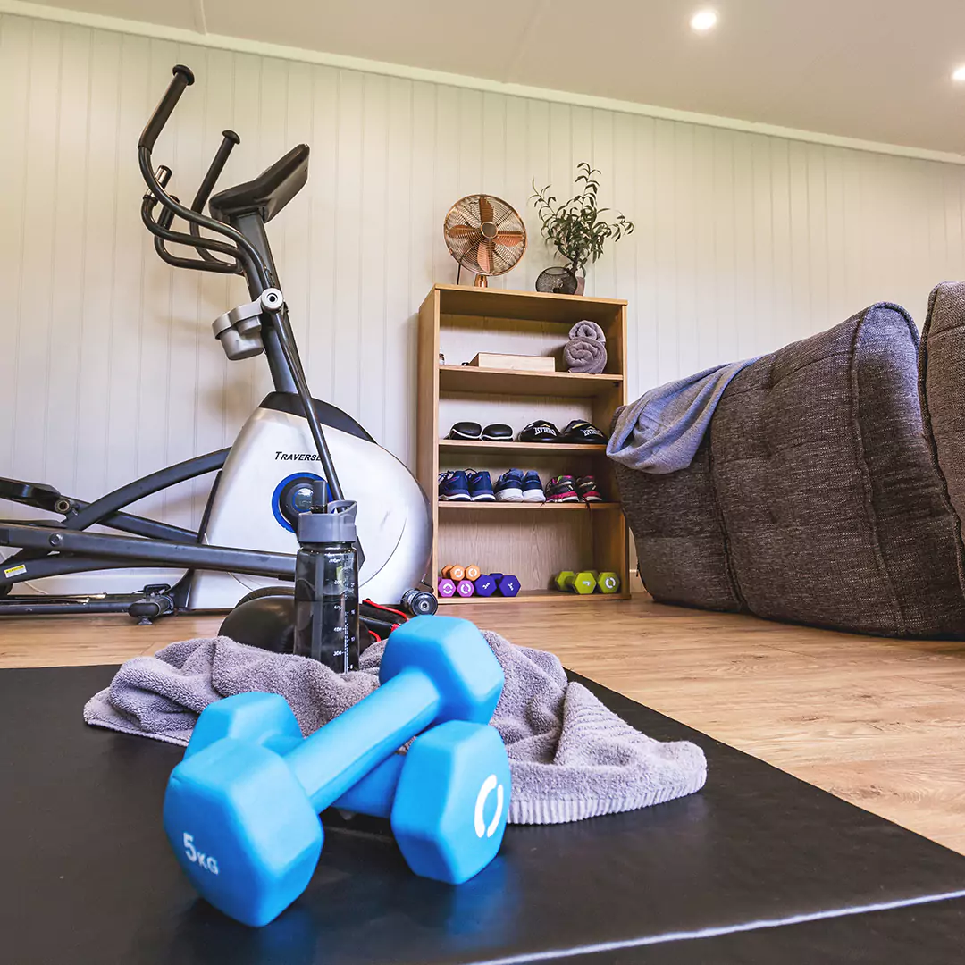 Cabin Master Garden Gym Building Internal shot with dumbbells in foreground and mat and towel and water bottle and dumbbell rack with elliptical machine and grey sofas