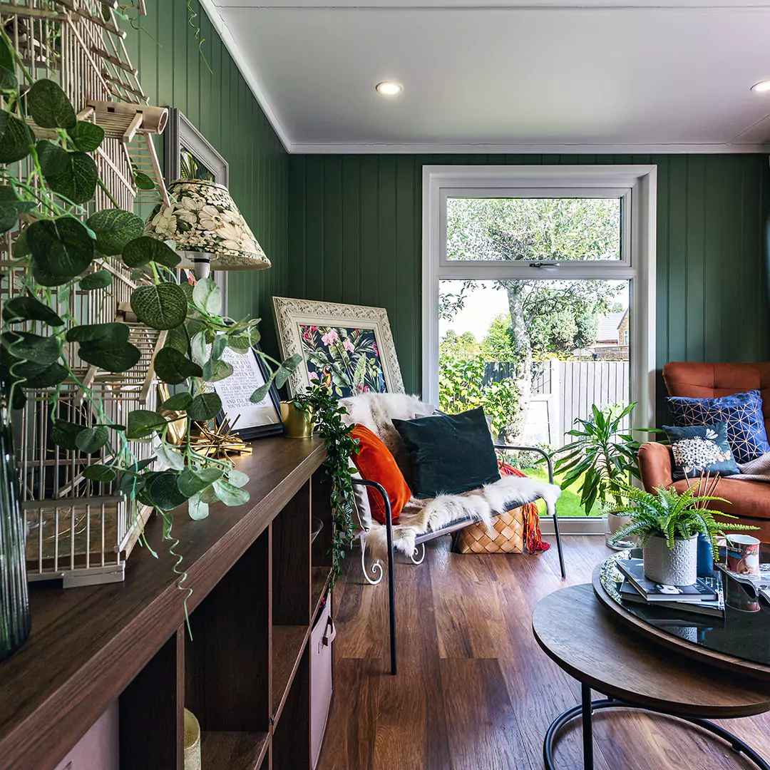 Interior of a garden room with dark green feature wall and green velvet sofa and green tropical plant