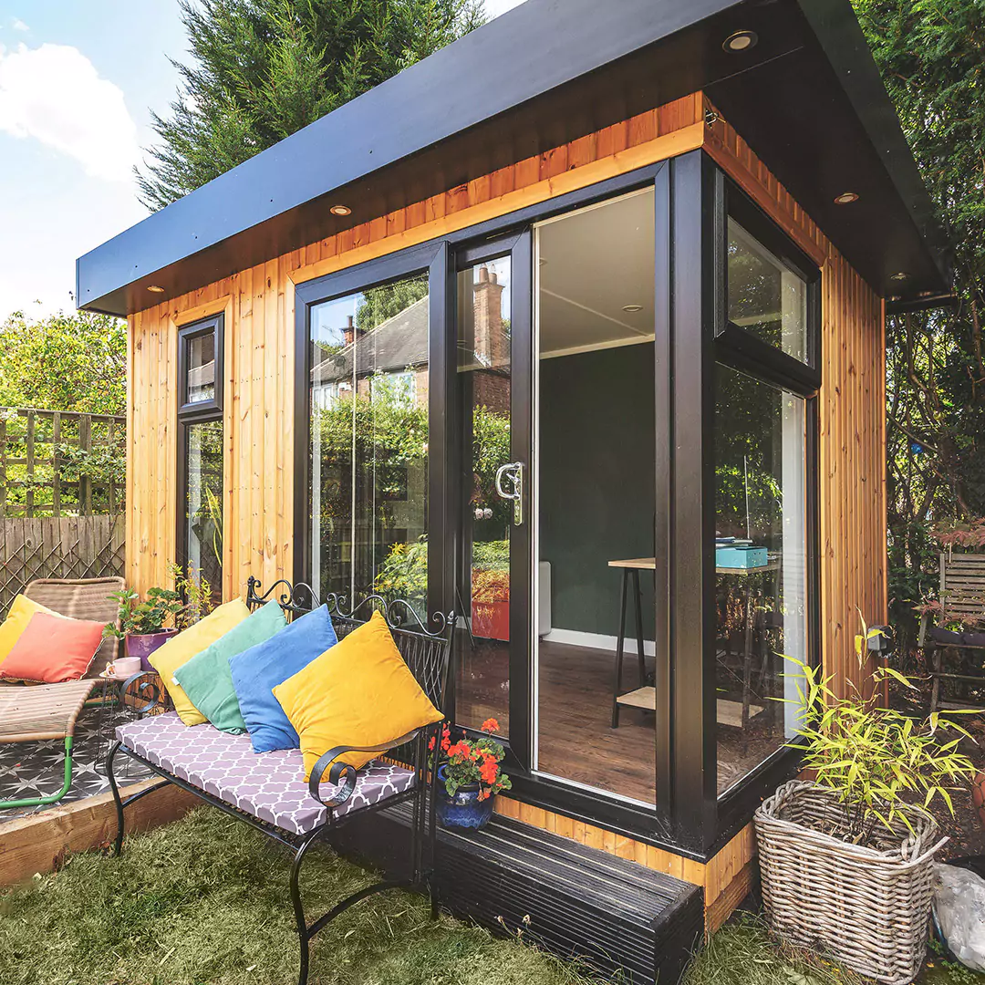 small garden office with bench and brightly coloured cushions with lawn area and potted plants 