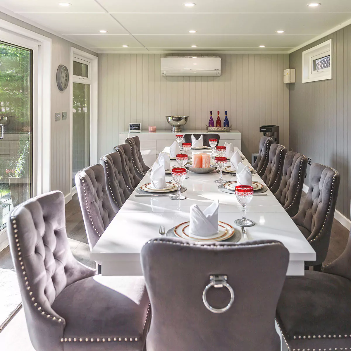 internal view of garden dining room with grey velvet chairs and large white dining table with plates and cutlery and drinks cabinet in background