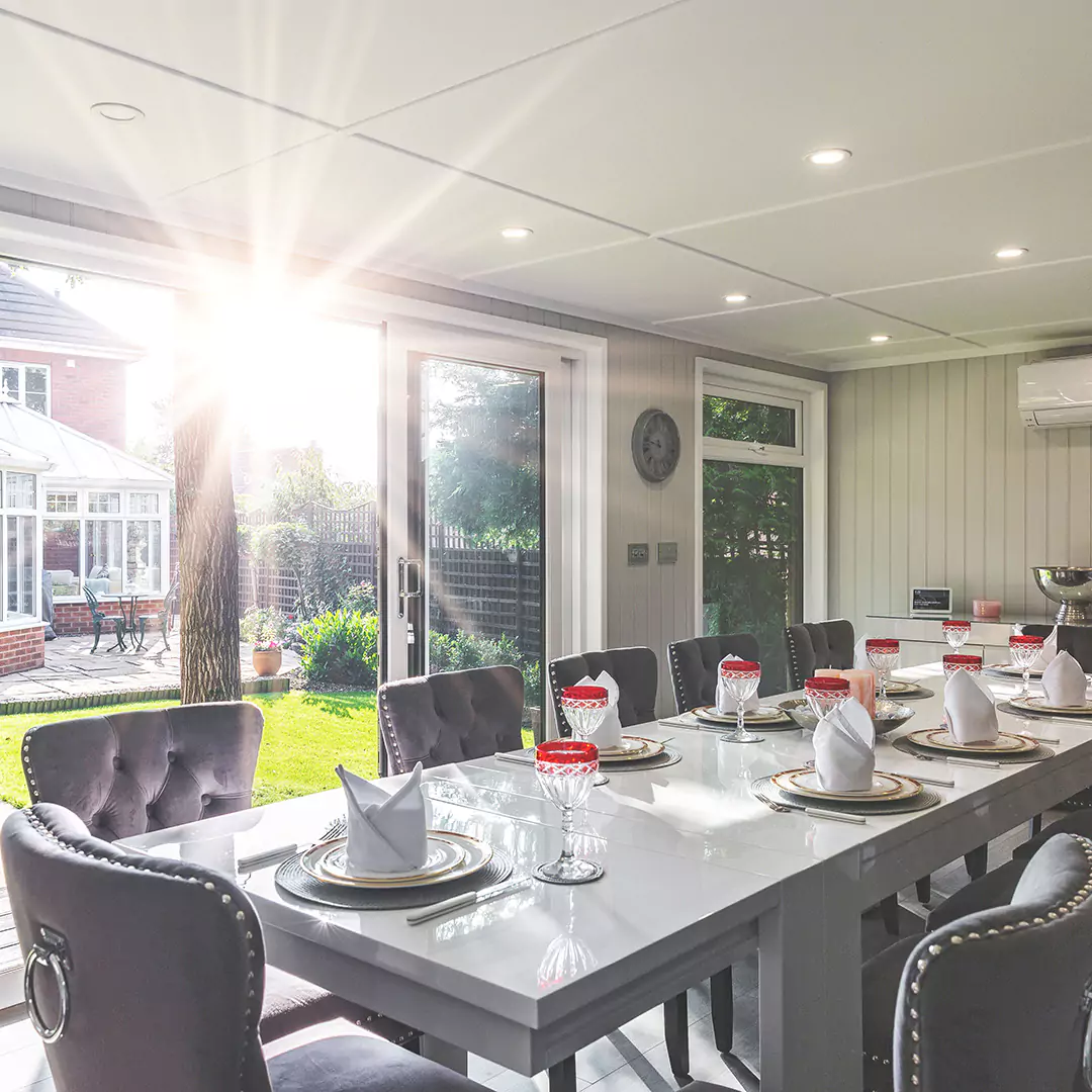 internal view of garden dining room with grey velvet chairs and large white dining table with plates and cutlery and drinks cabinet in background and view of large green lawn area with oak tree 