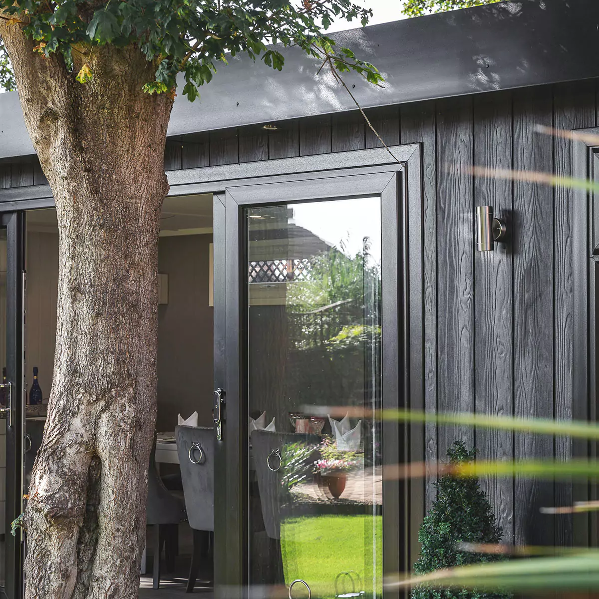 Zero maintenance Marley Garden Dining Room Cabin Master Timber Building with large oak tree in front of open sliding doors and potted plant at the side