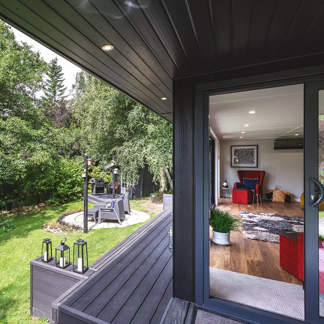 Black Marley Cabin Master Garden Building side profile with view of green lawn and rattan furniture and patio area with view into building with indoor plant and red velvet sofas and rug