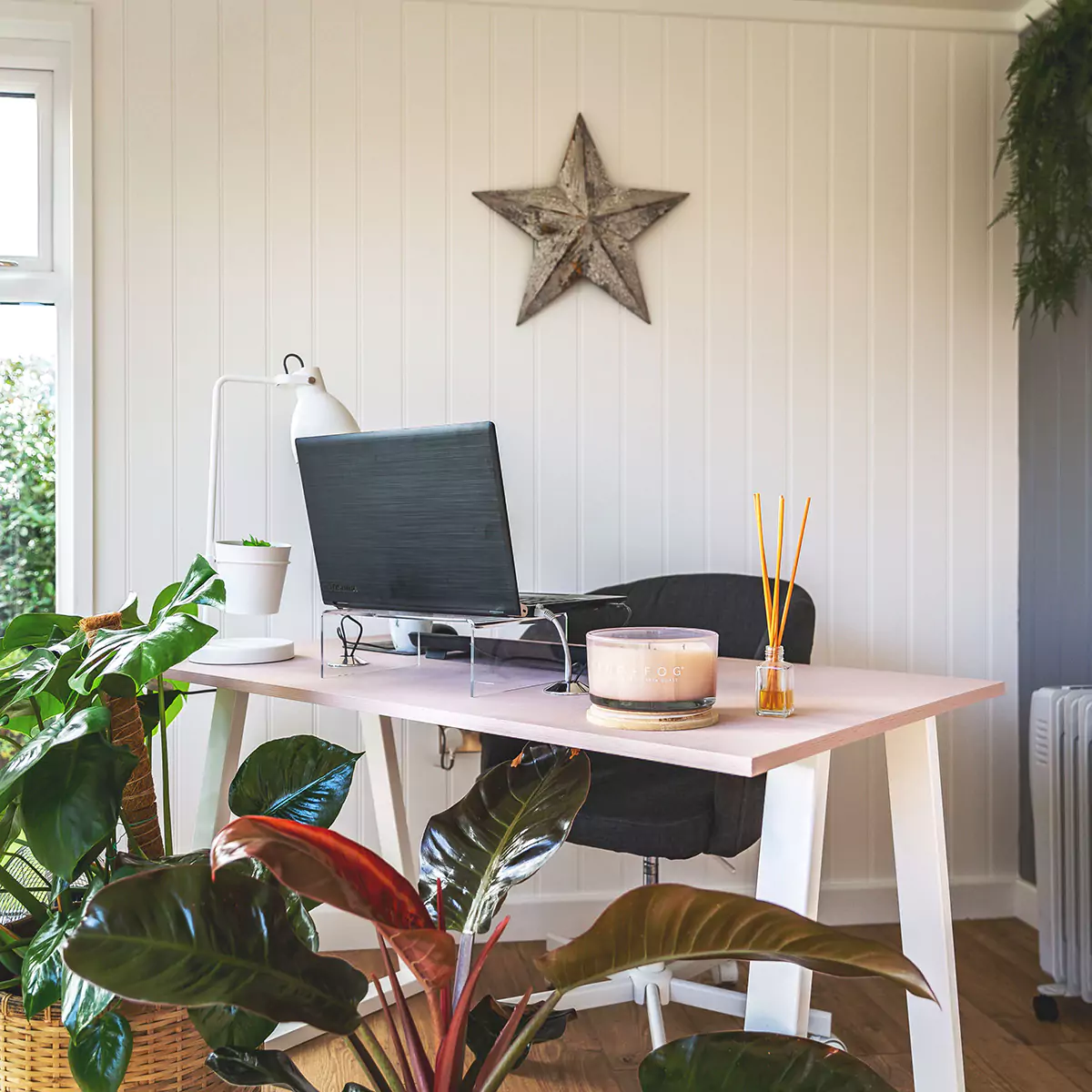 desk and chair with candles and diffuser on top of it, surrounded by indoor house plants inside garden office space with wall art and large grey feature wall