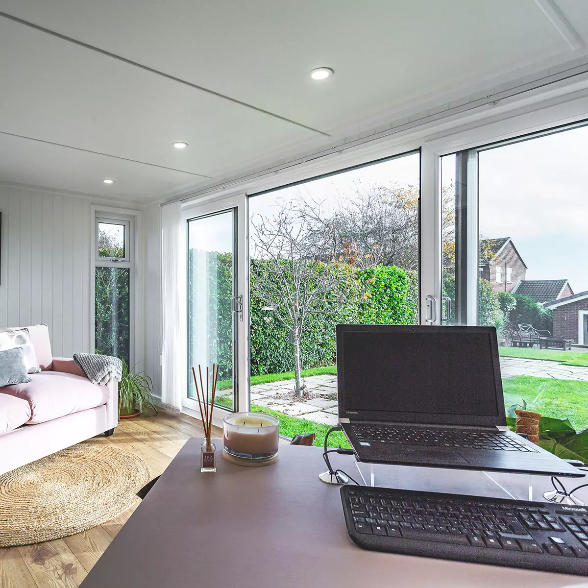 garden office space with sliding glass doors and desk with candle and diffuser and laptop on it with pink couch 